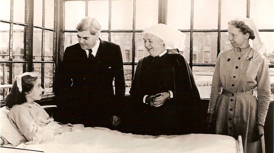 A black-and-white photo of a hospital ward. A young girl, one of the first NHS patients, lies in a bed, being visited by Nye Bevan and two nurses.